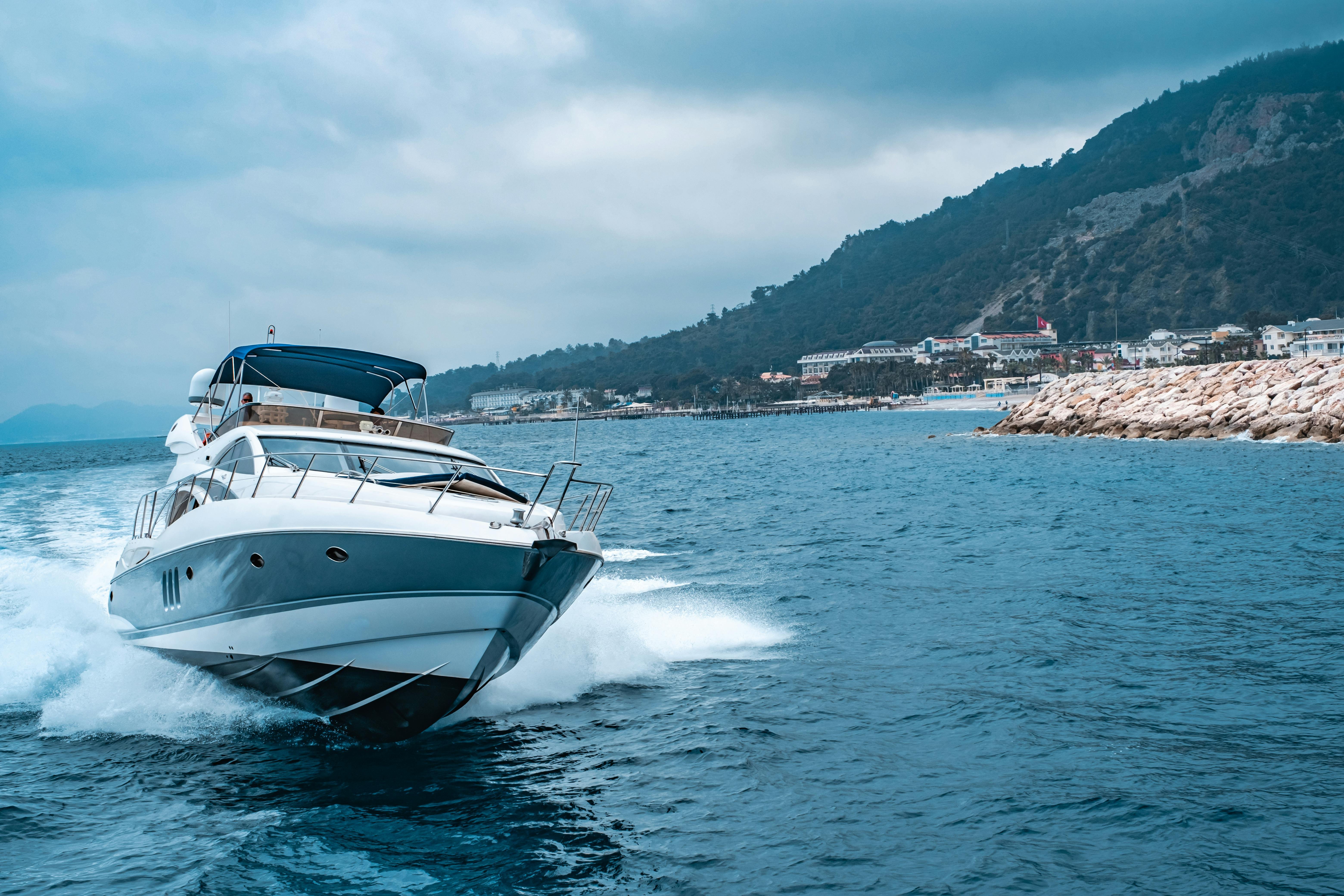 Bateau à moteur blanc et bleu naviguant vite près de la côte rocheuse, eau bleue et remous devant un littoral avec ville et montagnes en arrière-plan. Bateau à moteur blanc et bleu naviguant vite près de la côte rocheuse, eau bleue et remous devant un littoral avec ville et montagnes en arrière-plan.
