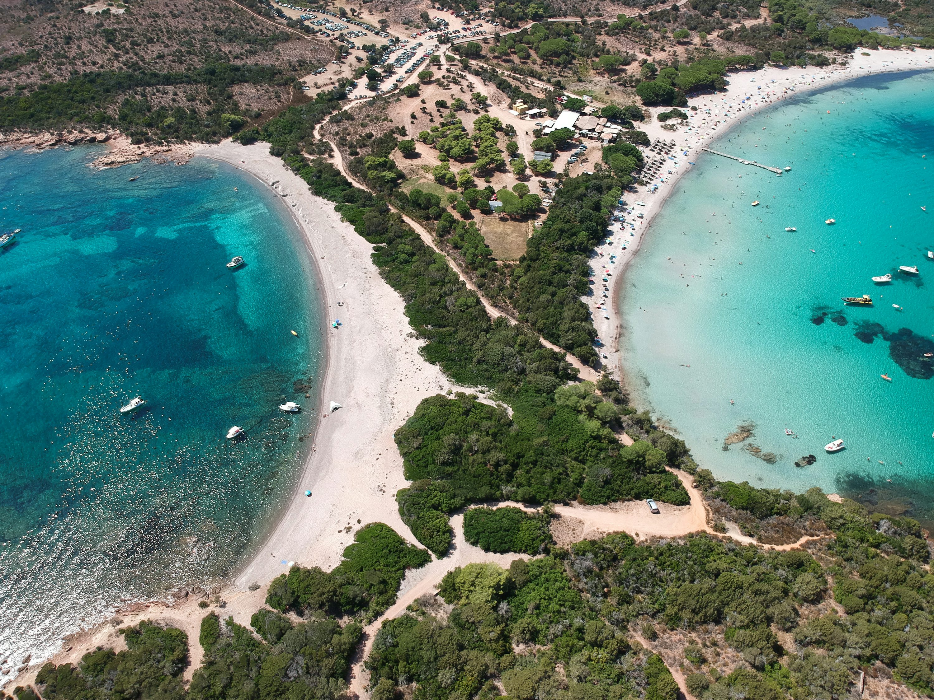 Vue aérienne d'une plage en forme de demi-lune entre deux eaux turquoise, entourée de forêts et de parasols sur le sable. Vue aérienne d'une plage en forme de demi-lune entre deux eaux turquoise, entourée de forêts et de parasols sur le sable.