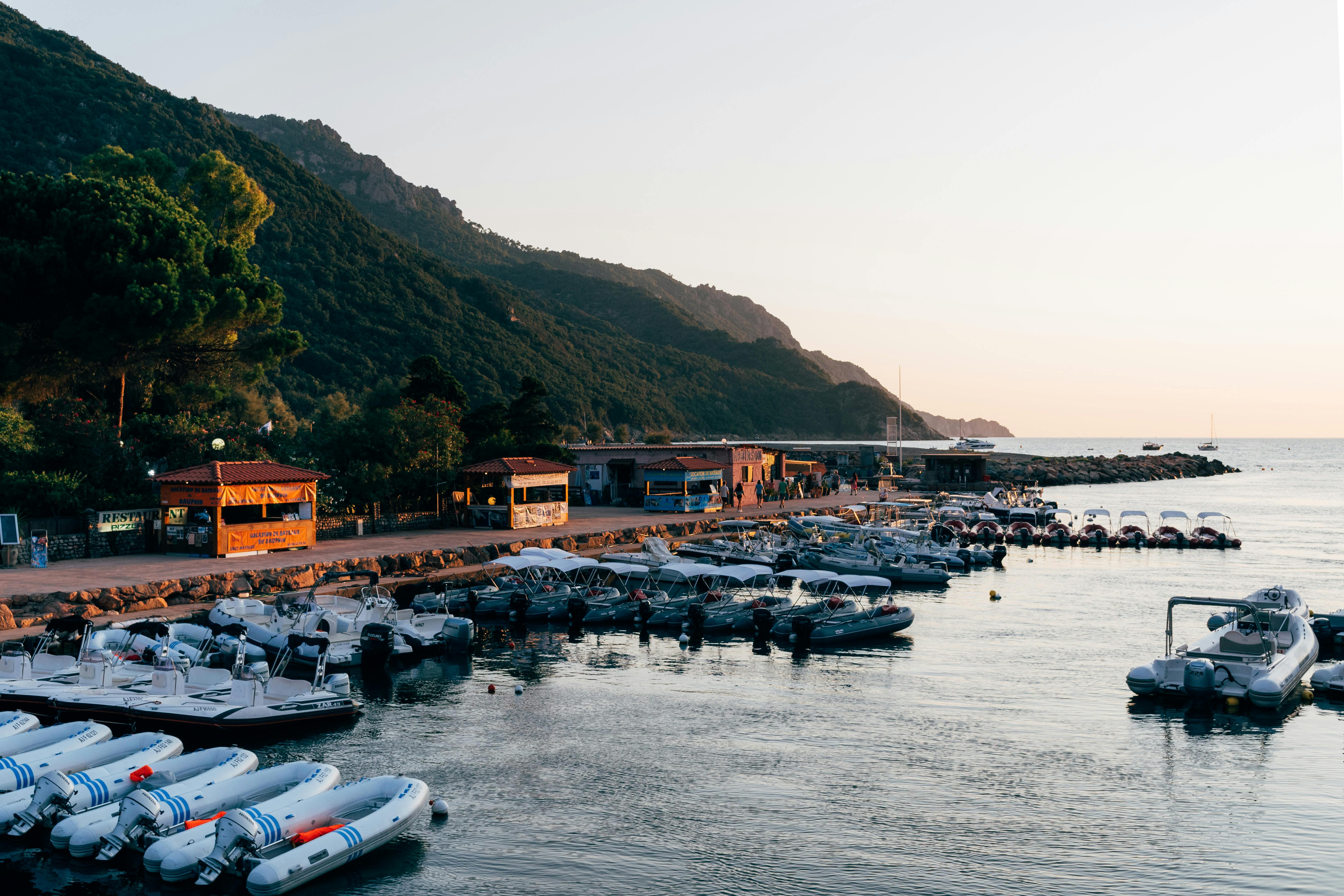 Port de plaisance animé au bord d’une côte rocheuse, avec bateaux et jet-skis amarrés, commerces et promeneurs au quai, et montagnes verdoyantes au loin. Port de plaisance animé au bord d’une côte rocheuse, avec bateaux et jet-skis amarrés, commerces et promeneurs au quai, et montagnes verdoyantes au loin.